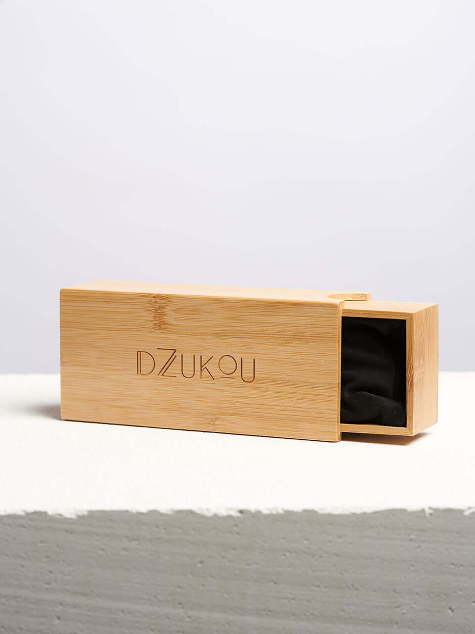 Bamboo sunglasses case with the Dzukou logo engraved on the front, showcasing a sliding drawer design with a black fabric lining inside, placed on a clean, textured surface.