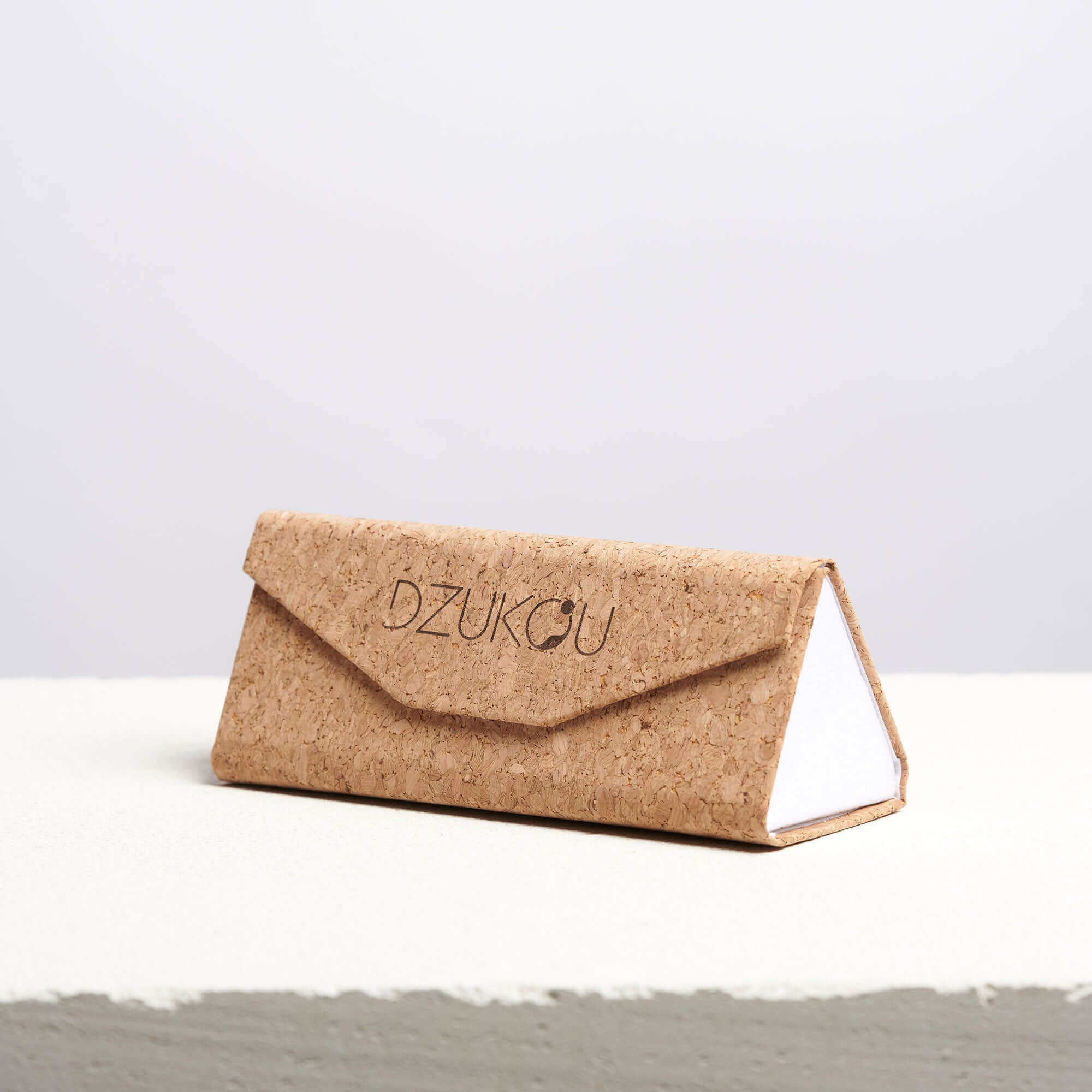 Eco-friendly cork sunglasses case with the Dzukou logo engraved on the front, featuring a triangular foldable design with a clean, white inner lining, displayed on a textured surface.