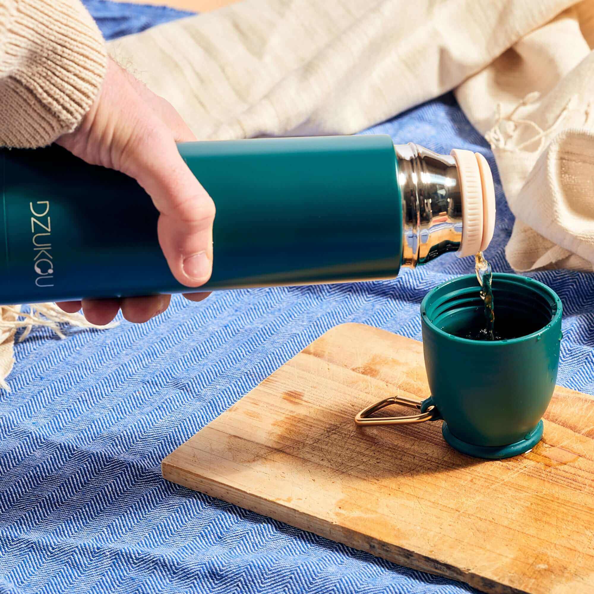 A person elegantly pours tea from a stainless steel thermos flask into a cup placed on a table, showcasing a serene tea moment.