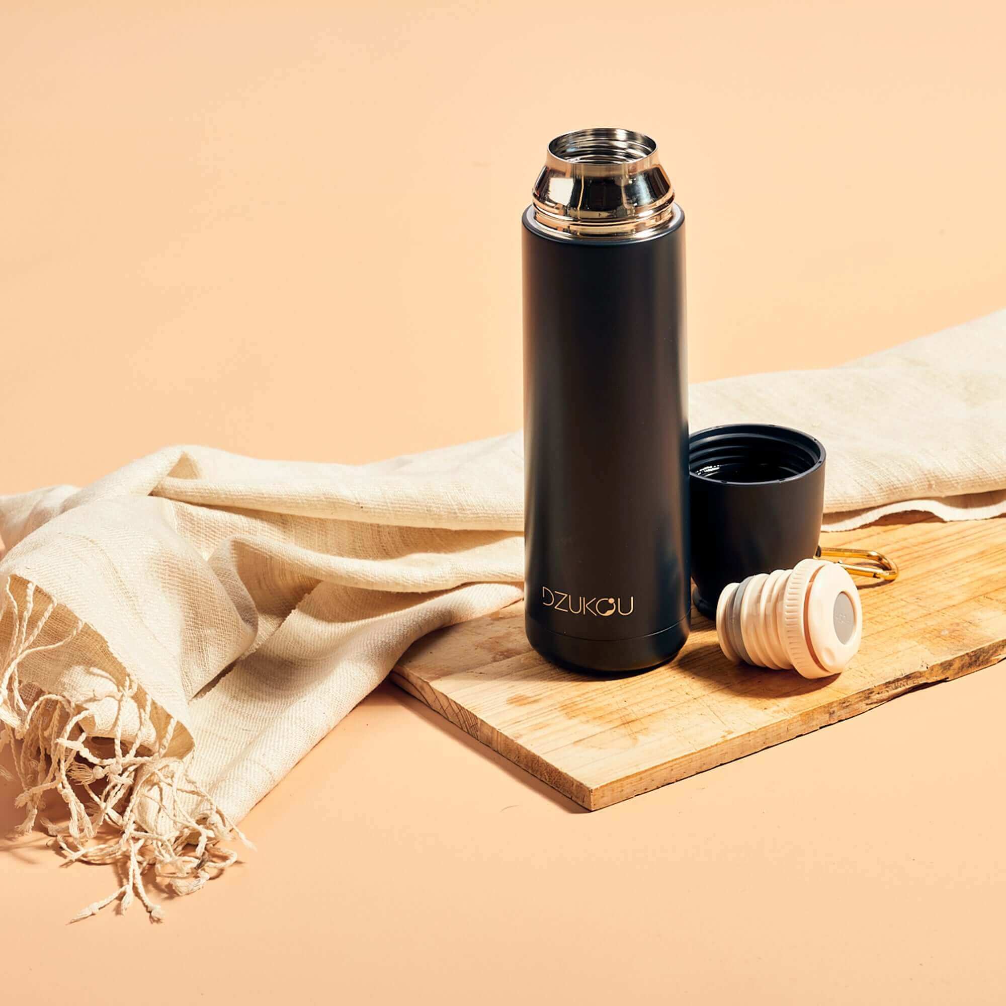 A stainless steel thermos flask and a cup placed on a wooden board with a eri silk stole in a background, suggesting a outdoor beverage setting.