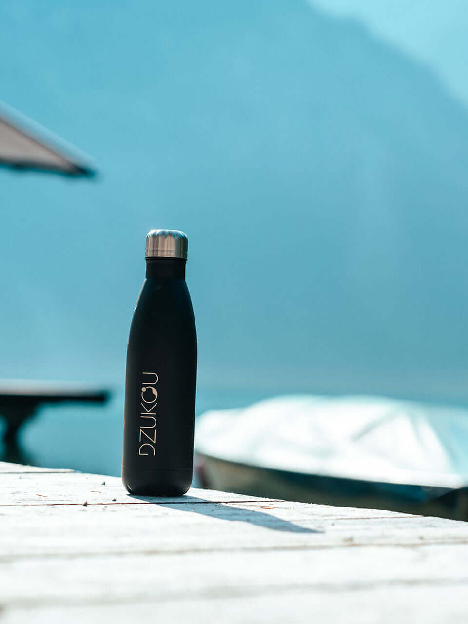 A black water bottle rests on a wooden table beside a serene lake, reflecting nature&