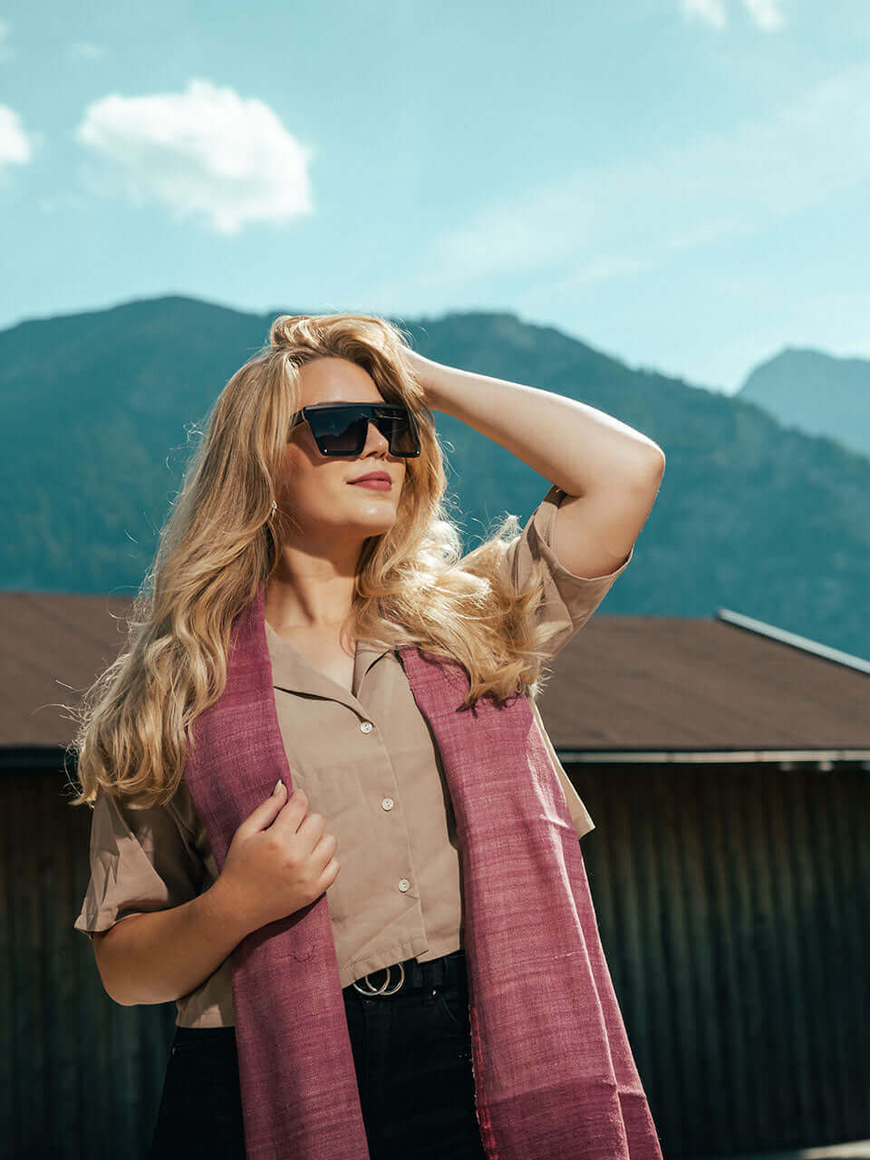 A woman wearing sunglasses and a pink scarf stands in front of a rustic cabin, exuding a sense of style and tranquility.
