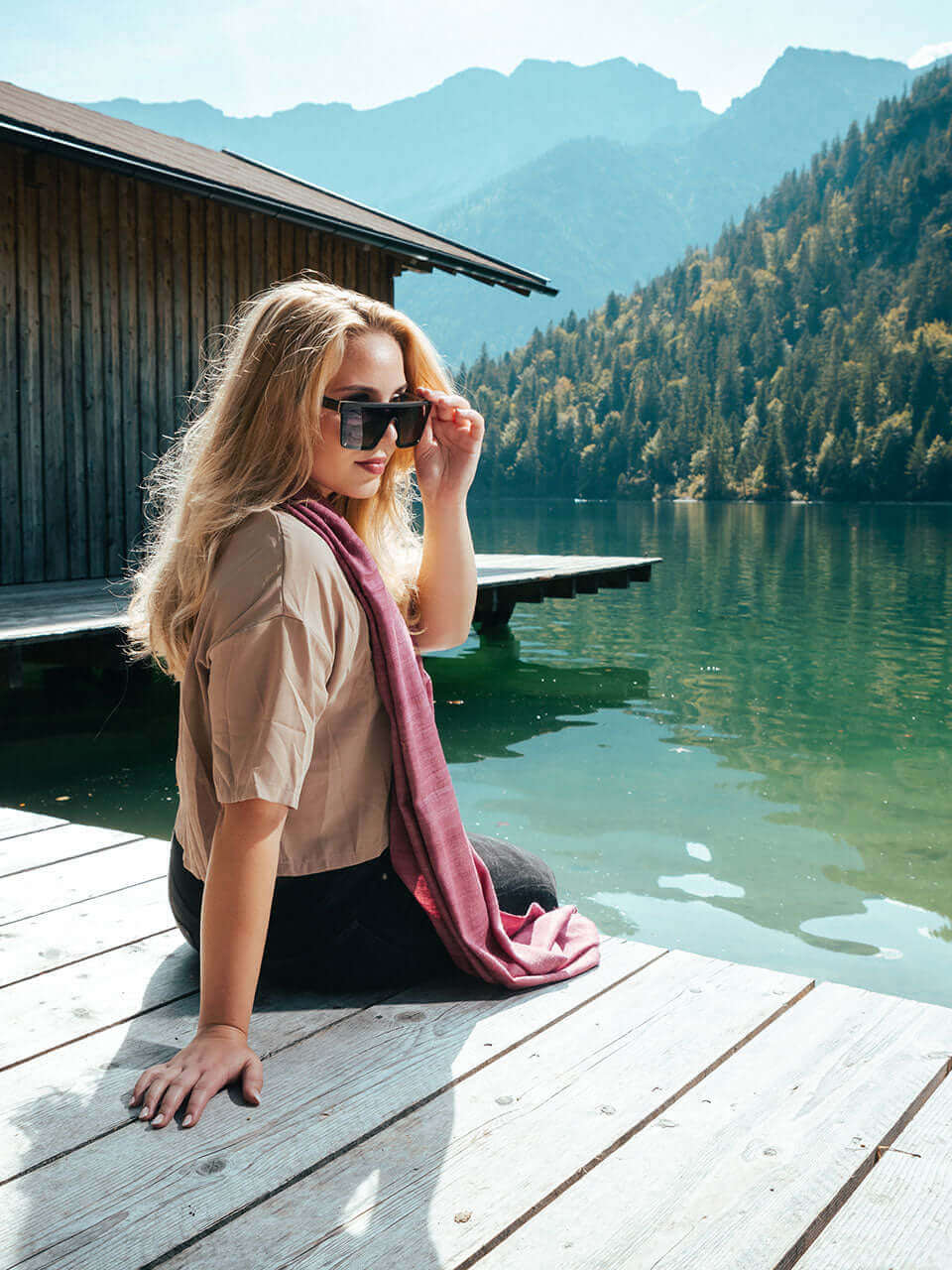 A woman seated on a dock wearing a pink scarf, gazing at the serene lake, surrounded by nature&