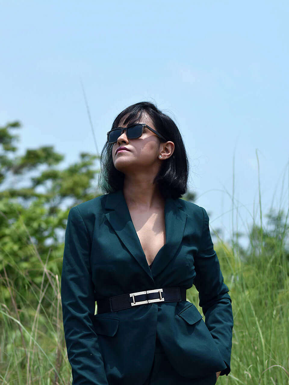 A woman in a green suit and wooden sunglasses stands confidently in a vibrant field, enjoying the sunny day.