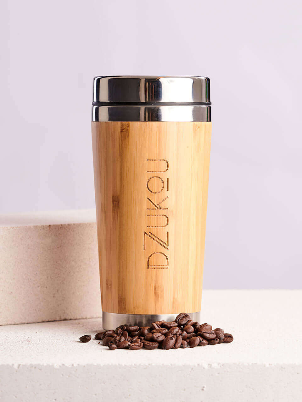 A bamboo coffee tumbler with a lid sits beside a scattered coffee beans on a textured surface.