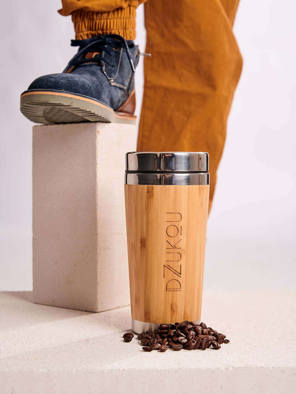 A person stands on a block surrounded by scattered coffee beans, and a bamboo coffee tumbler.
