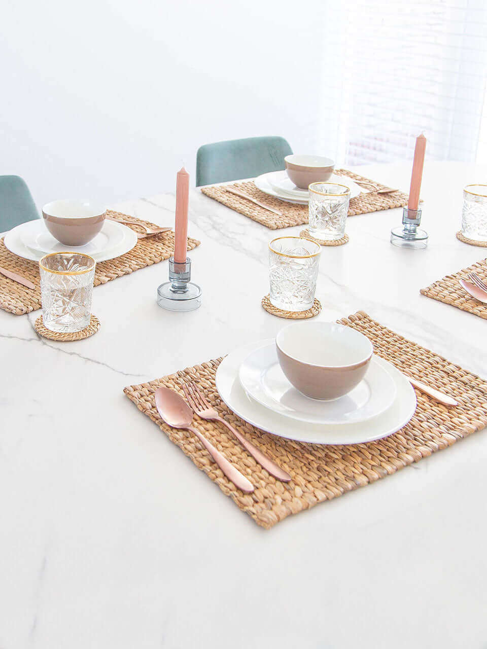 A side view of a table setting with a water hyacinth placemat, white ceramic dishes, rose gold cutlery, crystal glasses on cane coasters, all on a marble surface.