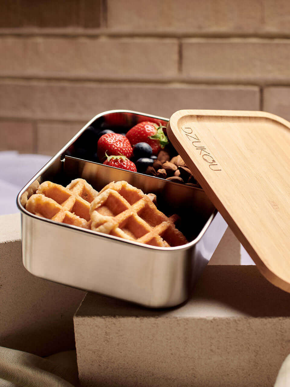 A bamboo lunch box containing waffles and assorted fresh fruit on top of a white block, arranged neatly for a balanced meal.