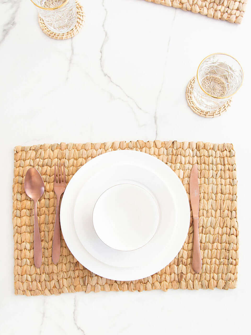 A table setting with a water hyacinth placemat, white ceramic dishes, rose gold cutlery, crystal glasses on cane coasters, all on a marble surface.