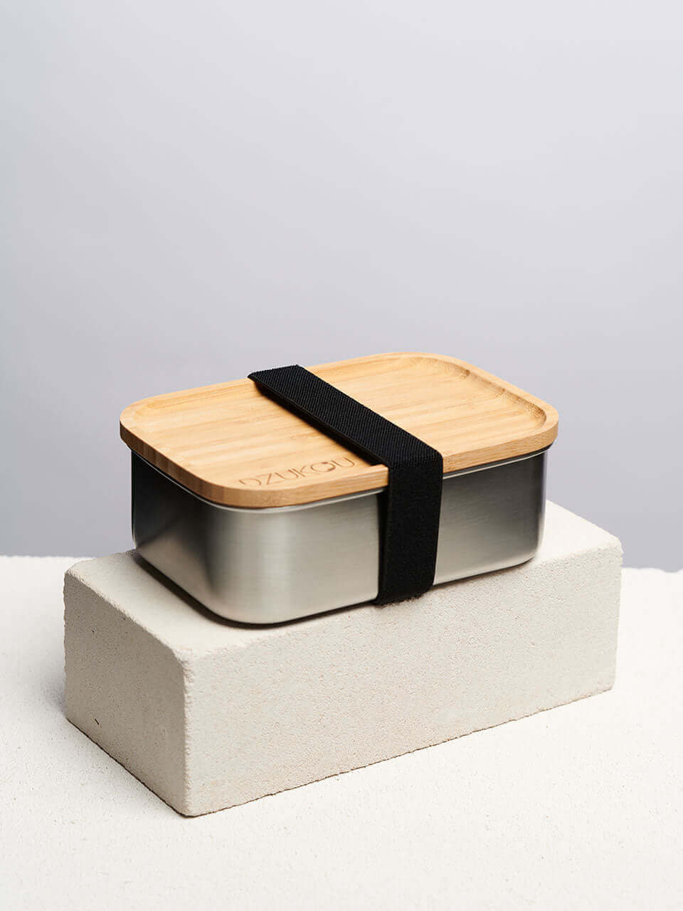 A bamboo lunch box secured with a black strap on top of a white block, showcasing a natural finish and a sleek, minimalist design.