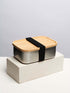 A bamboo lunch box secured with a black strap on top of a white block, showcasing a natural finish and a sleek, minimalist design.