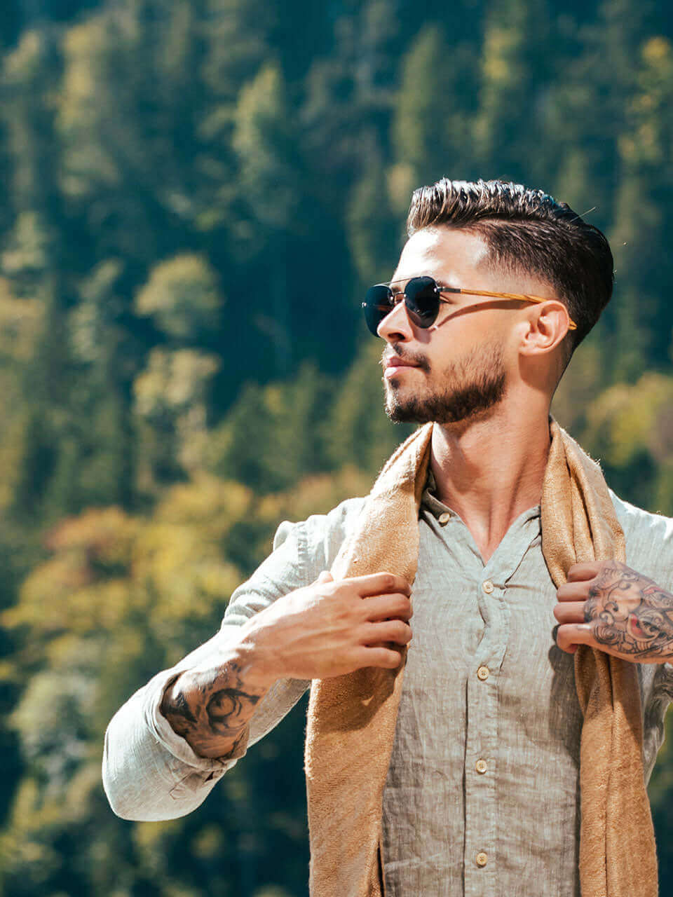 A tattooed man wearing a wooden sunglasses stands confidently amidst a serene forest backdrop.