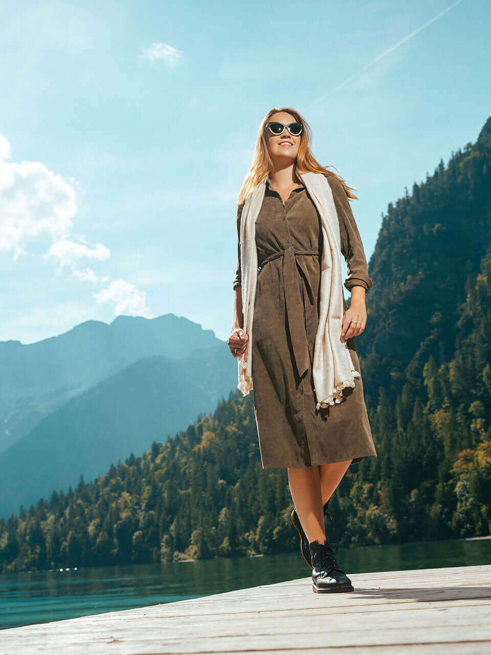 A woman wearing a dress and a cat eye wooden sunglass stands confidently on a dock, enjoying the scenic view.