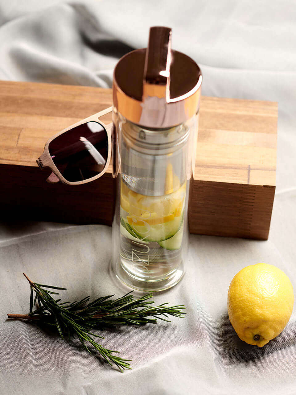 A glass bottle featuring lemon slices, cucumber, and a sprig of rosemary with a sunglass in back and a lemon on the sides, perfect for a summer drink.