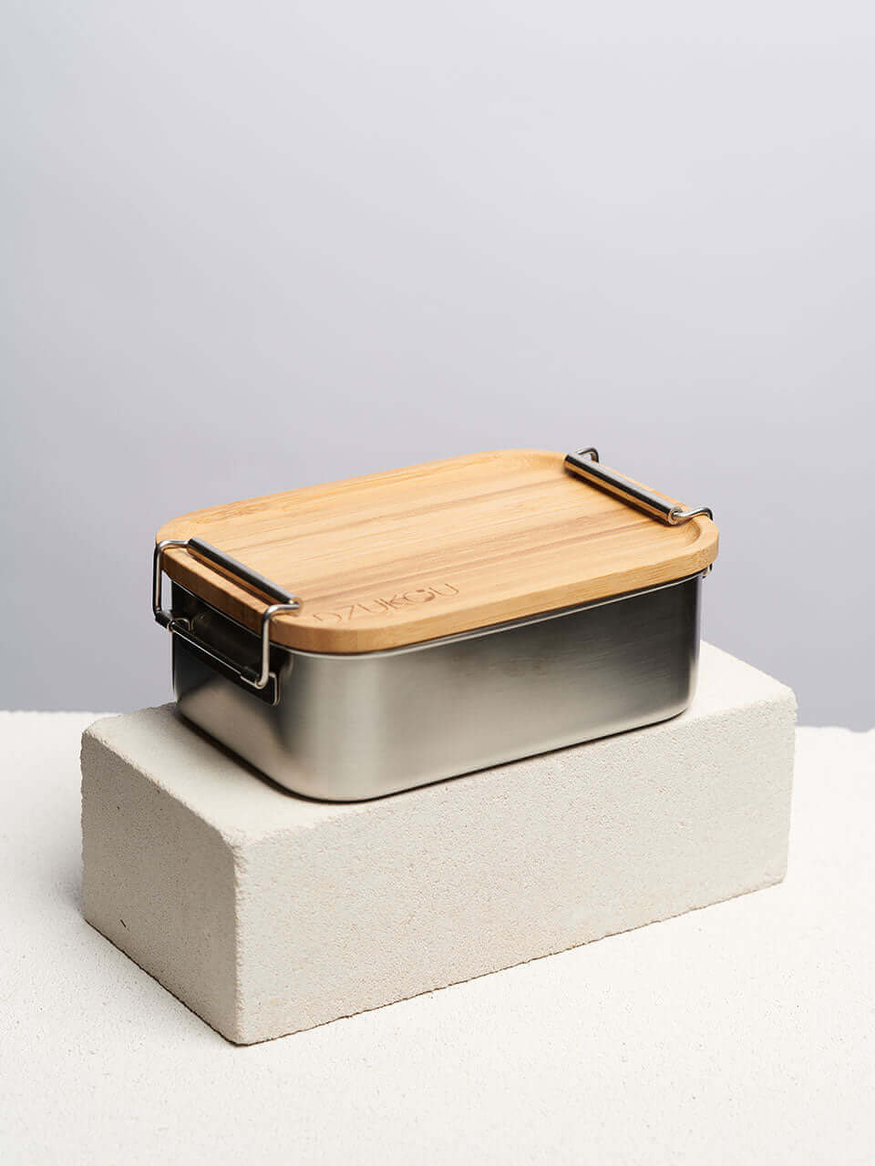 A bamboo lunch box secured with a black strap on top of a white block, showcasing a natural finish and a sleek, minimalist design.