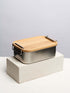 A bamboo lunch box secured with a black strap on top of a white block, showcasing a natural finish and a sleek, minimalist design.
