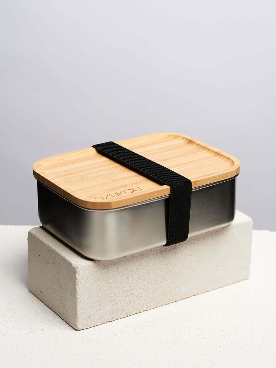 A bamboo lunch box secured with a black strap on top of a white block, showcasing a natural finish and a sleek, minimalist design.