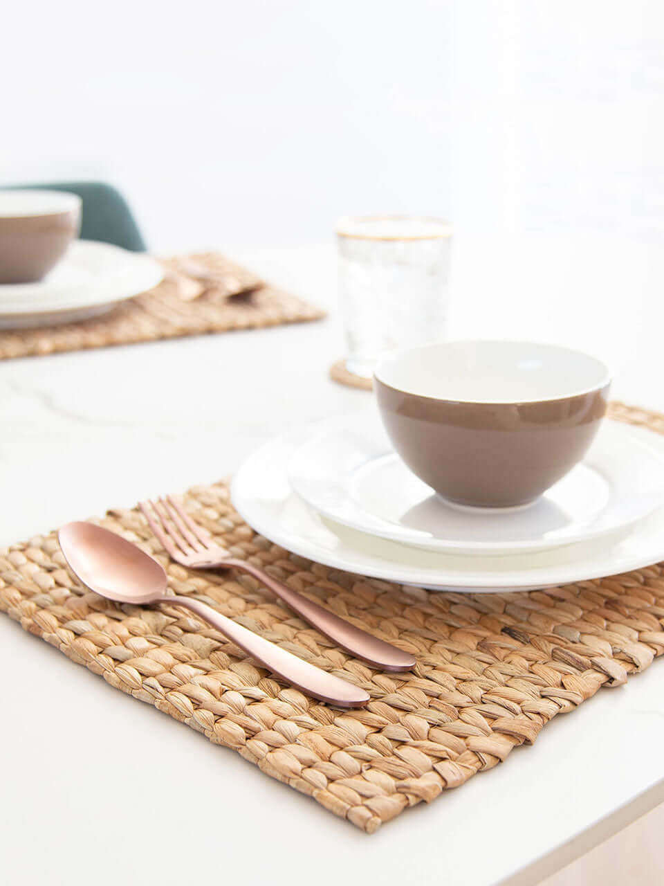 A side view of a table setting with a water hyacinth placemat, white ceramic dishes, rose gold cutlery, crystal glasses on cane coasters, all on a marble surface.