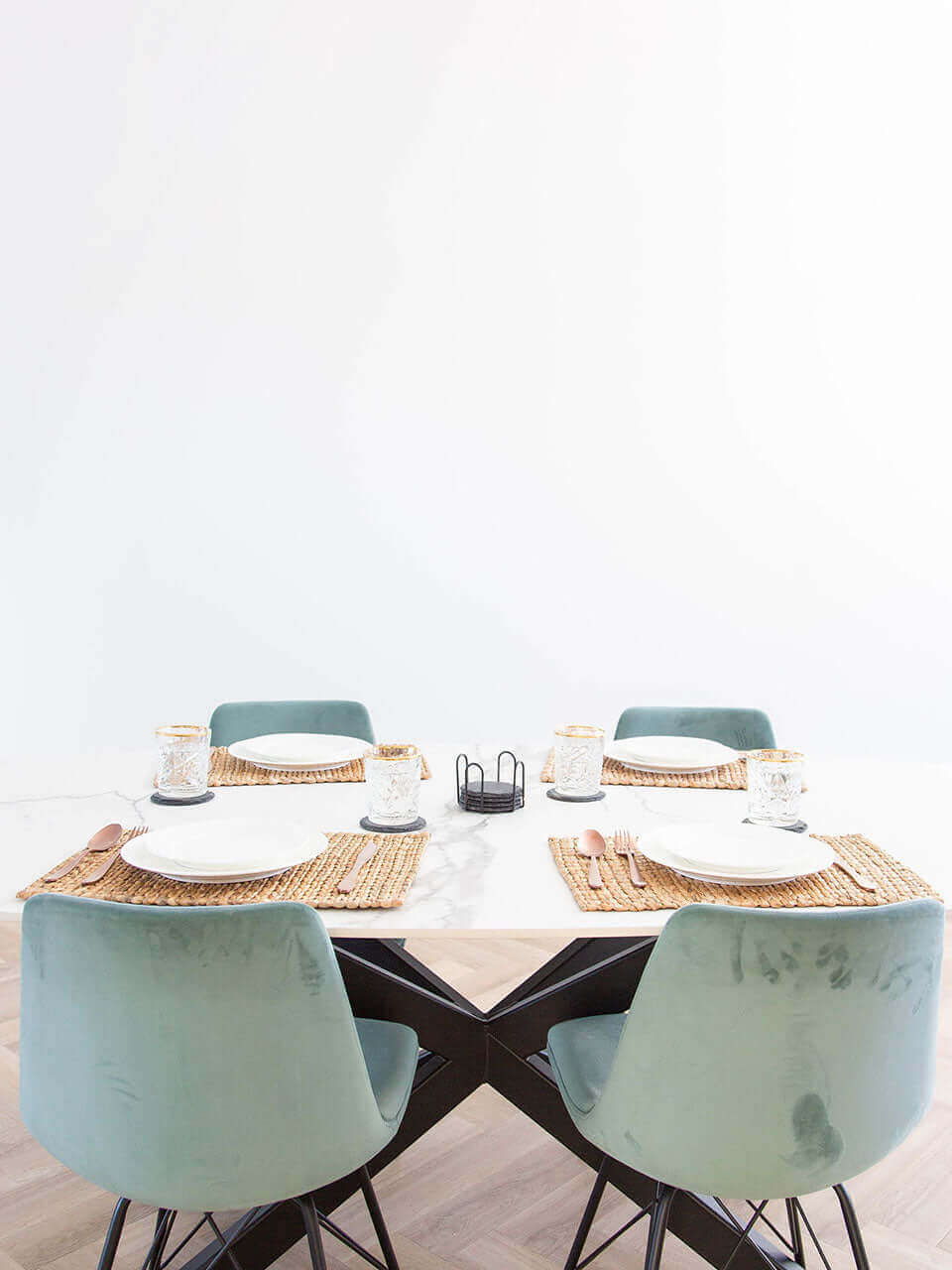 A dining table with four chairs, set with placemats, plates, cutlery, and glasses against a white wall.