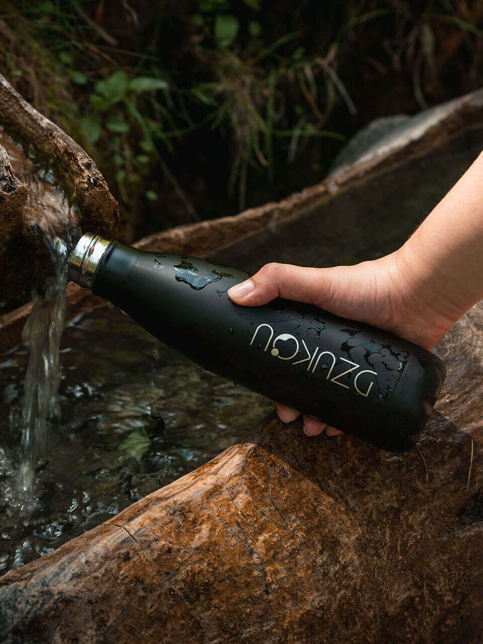 A person holds a black water bottle above a flowing stream, capturing the essence of nature and hydration.