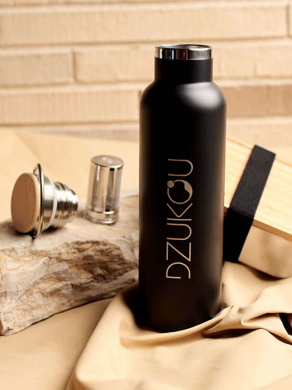 A stainless steel water bottle displayed against a beige background with a lunchbox behind and the lid and infuser on the side, highlighting its eco-friendly design.