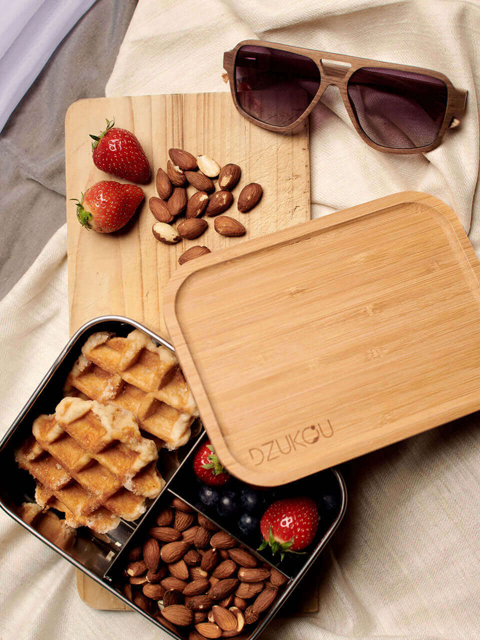 A bamboo lunchbox featuring waffles, strawberries, and assorted food items, accompanied by a pair of wooden sunglasses.