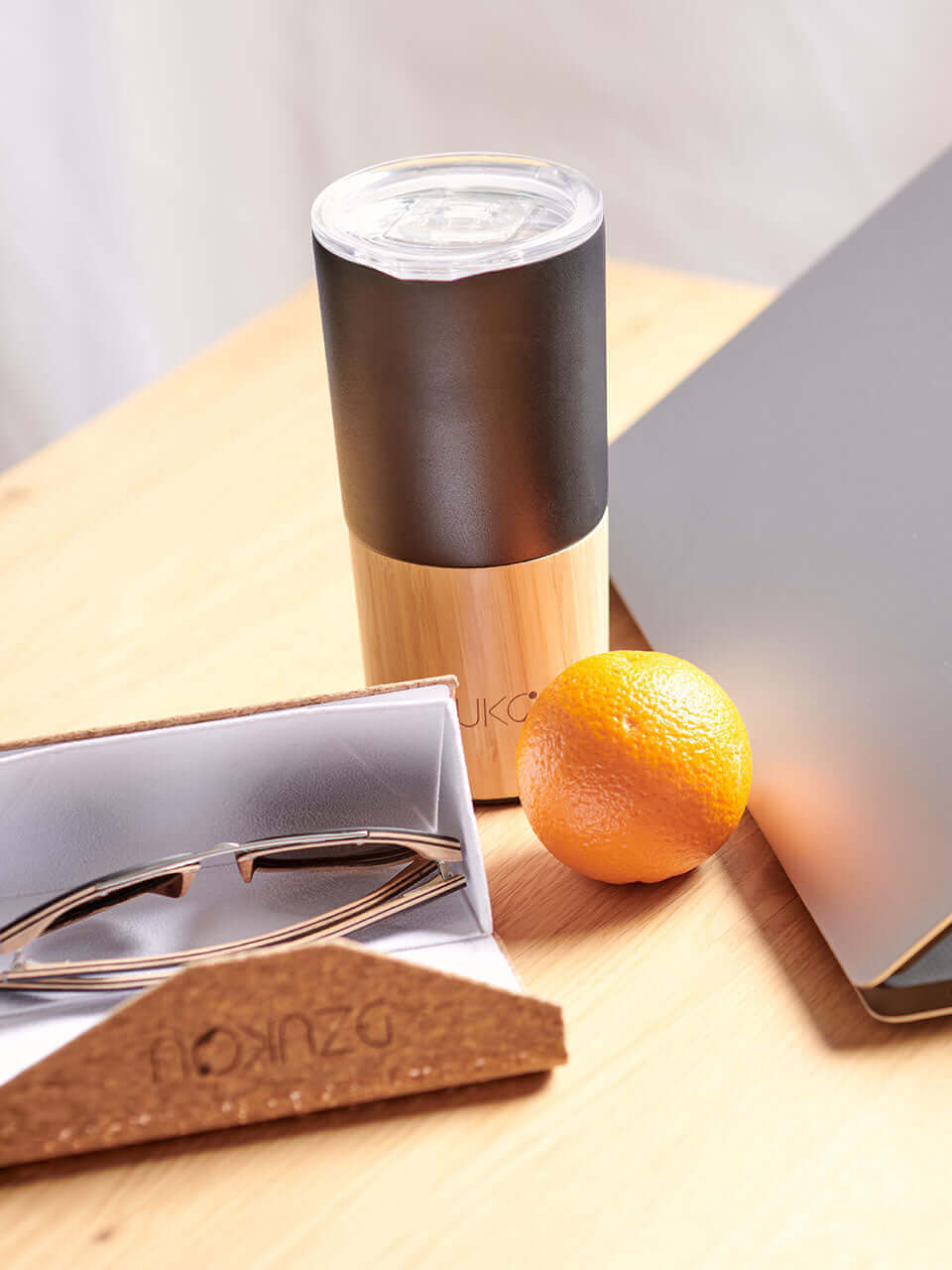 A black and bamboo coffee tumbler along with a laptop, a box, and an orange are arranged neatly on a wooden table, creating a simple yet organized workspace.