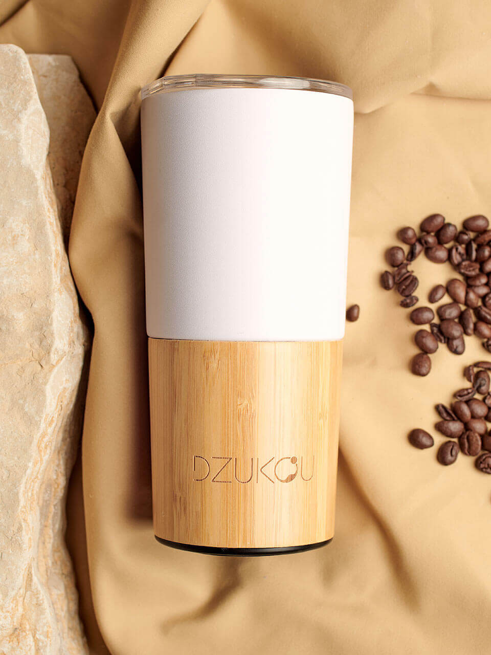 A bamboo coffee tumbler with a white lid, accompanied by scattered coffee beans on a beige background.