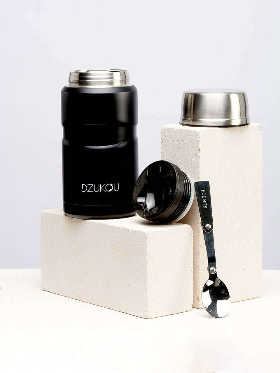 A black lunchpot with a lid and a spoon placed beside it, suitable for storing food or ingredients.