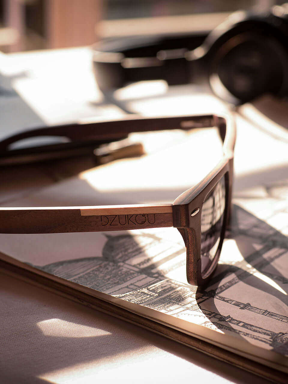 A wooden sunglass rests atop a book on a wooden table, suggesting a leisurely reading experience outdoors.
