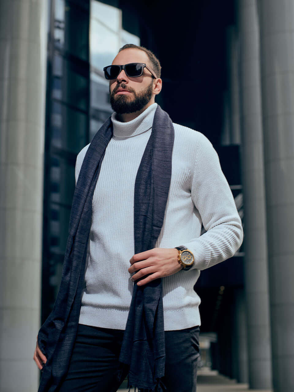 A man wearing a white sweater and grey scarf stands outdoors, surrounded by a serene environment.
