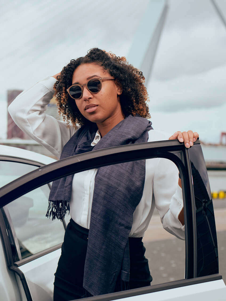 A woman wearing sunglasses and a grey scarf leans out of a car, enjoying the view with a confident expression.