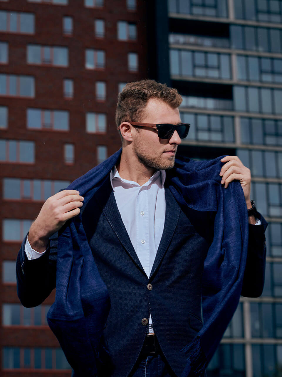 A man wearing a blazer and sunglasses along with a scarf stands confidently, exuding an air of mystery and sophistication.
