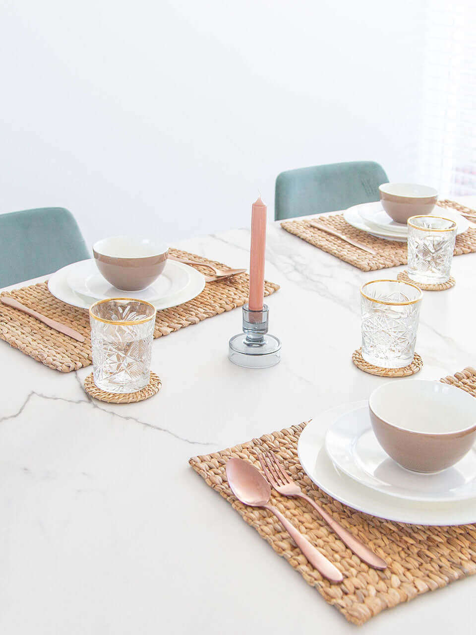 A side view of a table setting with a water hyacinth placemat, white ceramic dishes, rose gold cutlery, crystal glasses on cane coasters, all on a marble surface.