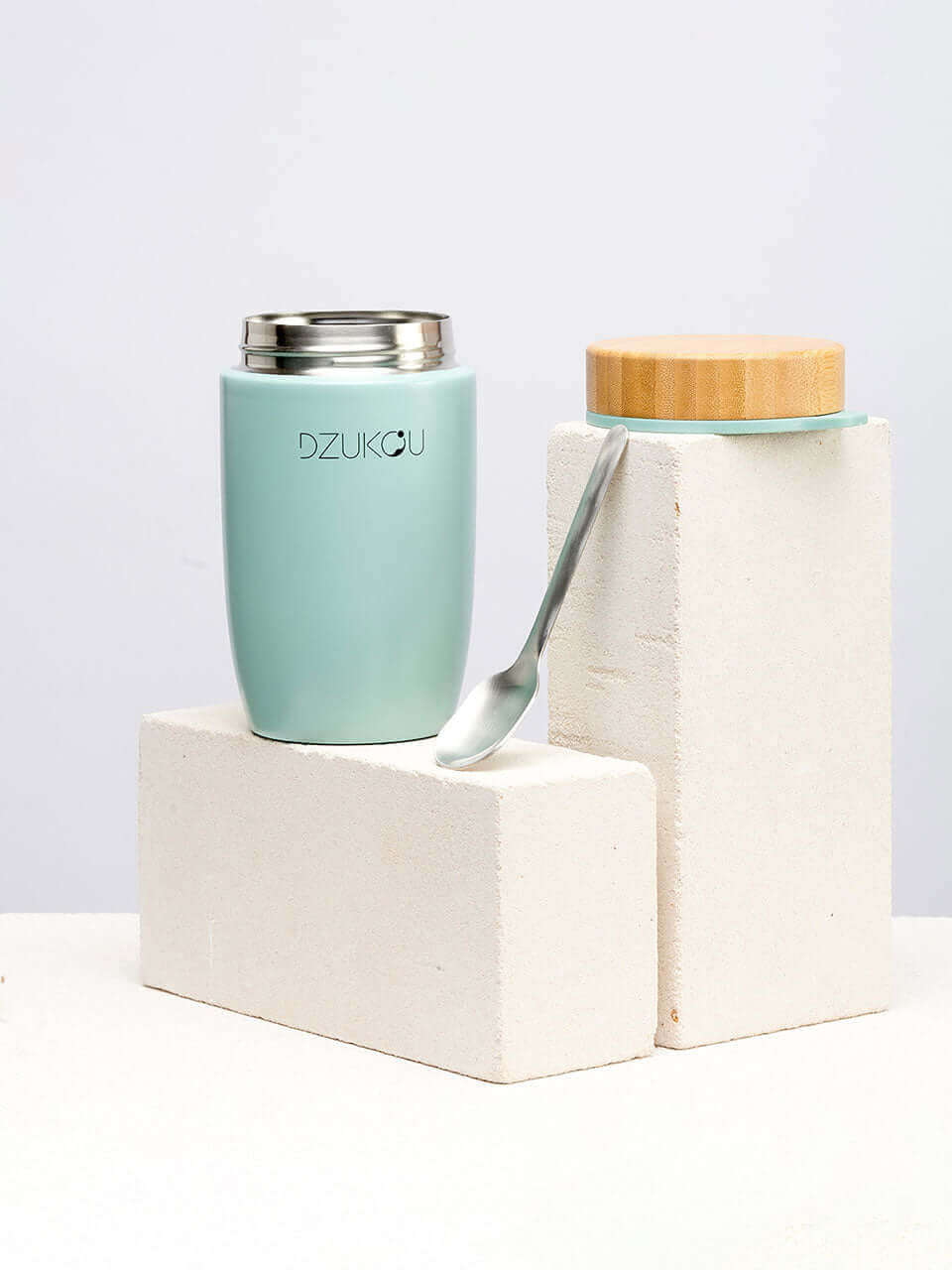 A bamboo green stainless steel lunchpot with a spoon resting beside it, topped with a wooden lid, elegantly displayed on a neutral surface.