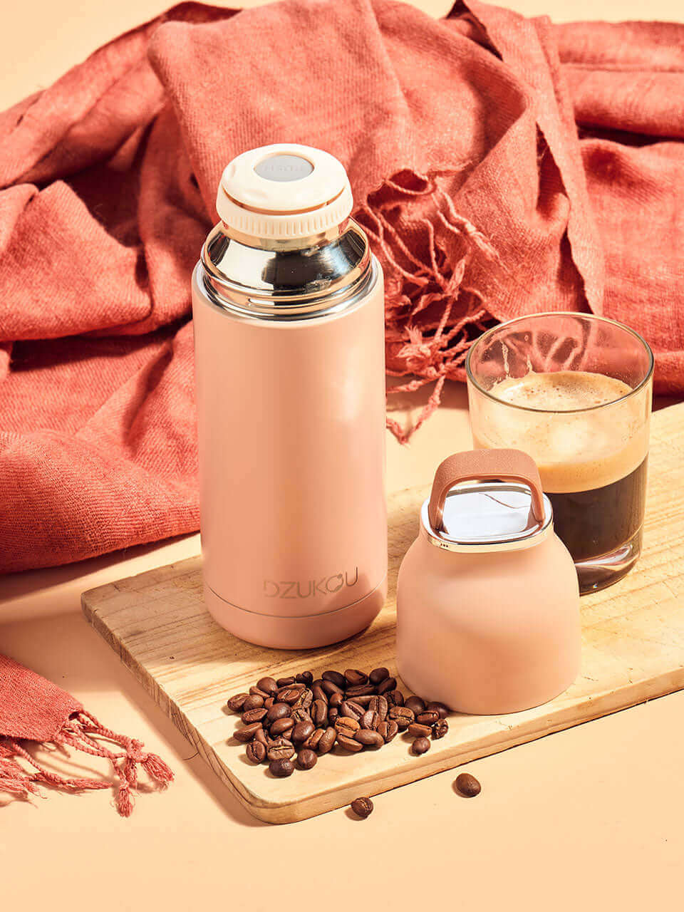 A peach stainless steel thermos bottle placed on a wooden cutting board surrounded by scattered coffee beans and a pink eri stole behind.