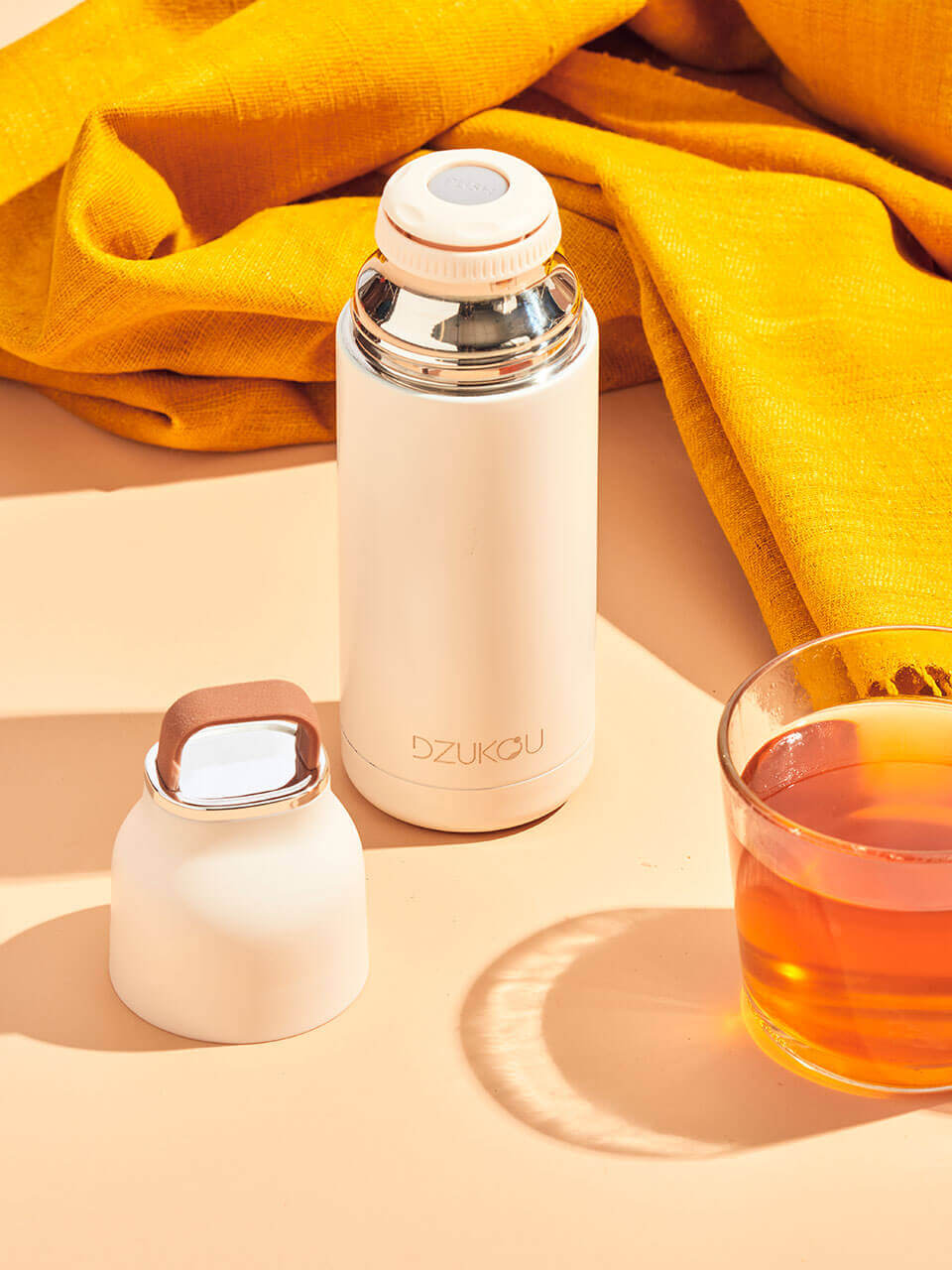 A white thermos bottle beside a steaming cup of tea on a wooden surface with a yellow eri silk stole behind, evoking warmth and comfort.