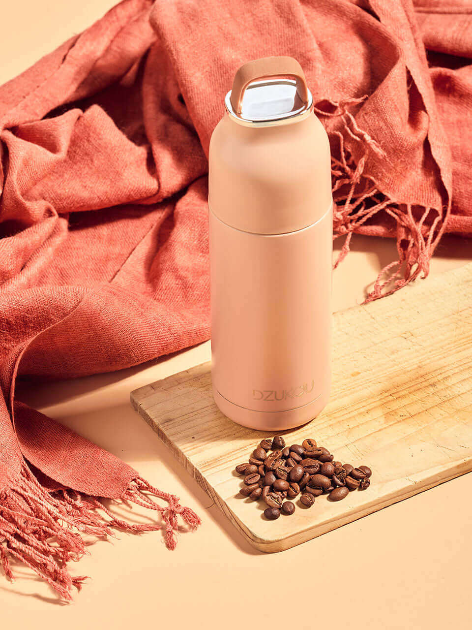 A peach stainless steel thermos bottle placed on a wooden cutting board surrounded by scattered coffee beans and a pink eri stole behind.