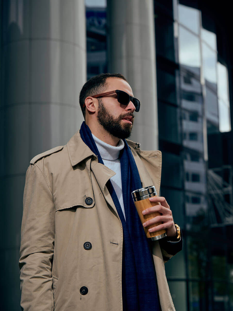 A man in a trench coat and eri silk scarf stands holding a bamboo coffee tumbler, exuding a casual yet stylish demeanor.