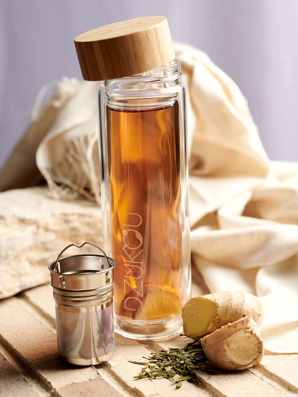 A glass bottle with a lid containing tea along with a infuser and some herbs on the sides, showcasing a natural and aromatic display.