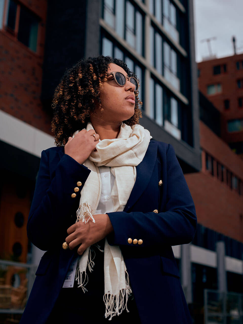 A woman in a black dress and scarf stands elegantly in front of a building, showcasing a poised and stylish demeanor.