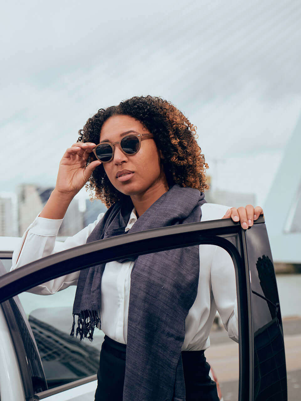 A woman in wooden sunglasses casually leans against the door of a car, exuding confidence and style.