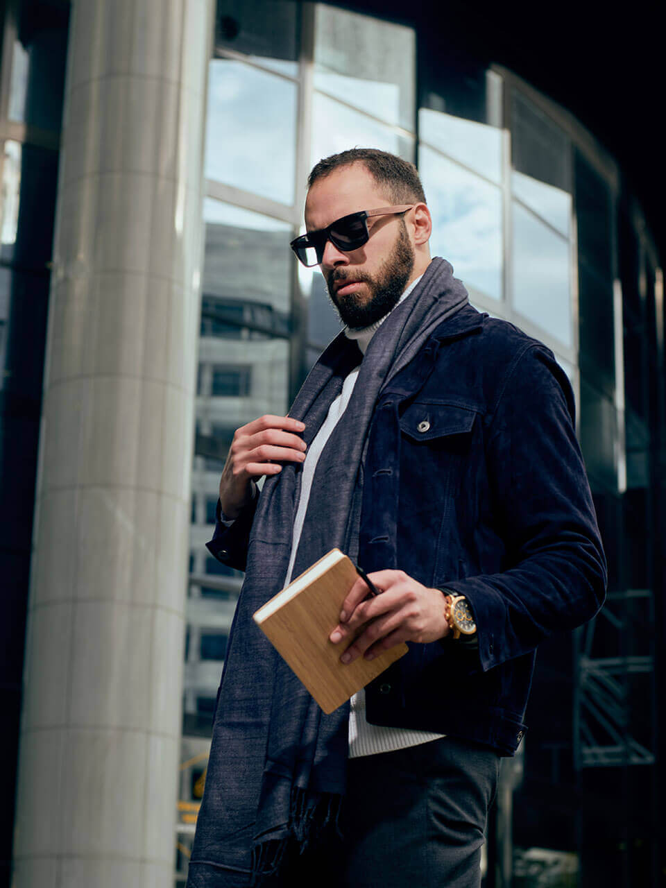 A man wearing a jacket , scarf and a wooden sunglasses is holding a notebook, appearing engaged and thoughtful.