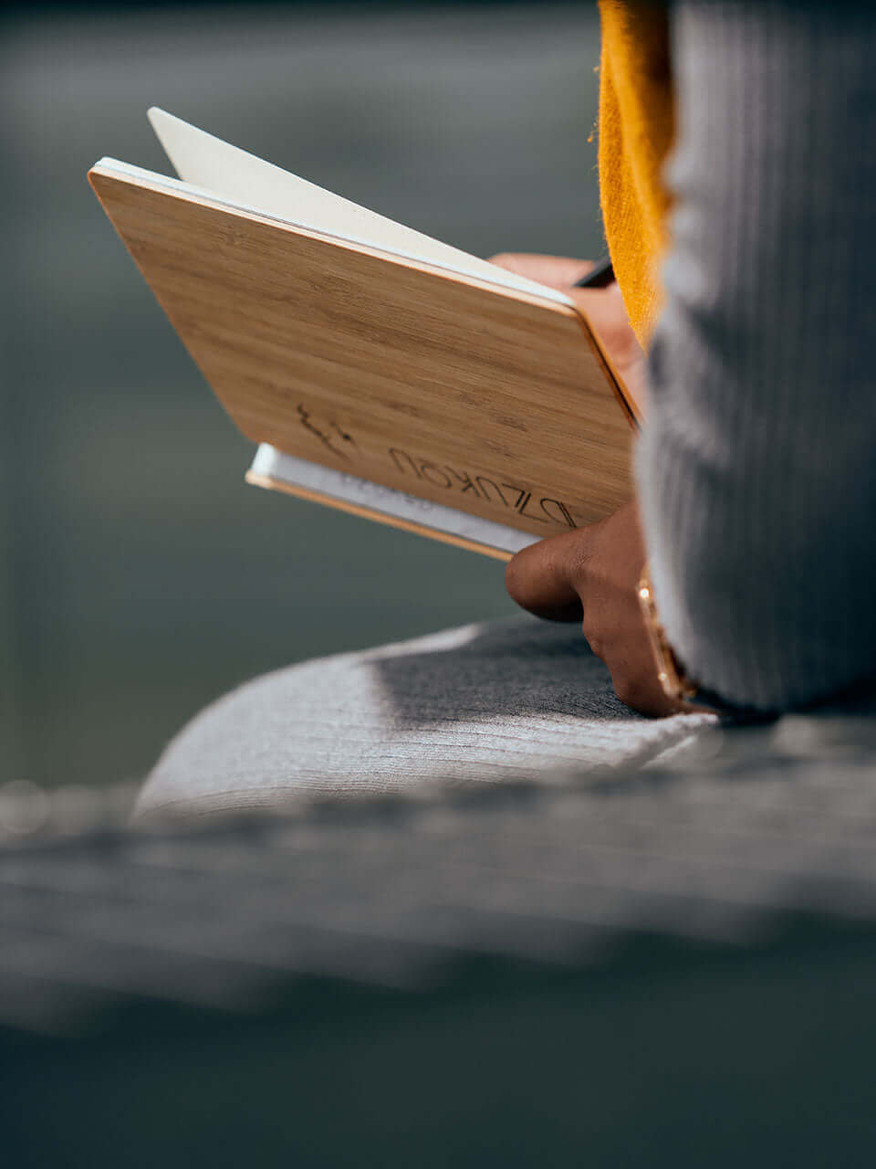 A person seated on a bench, engrossed in reading a bamboo notebook, surrounded by a serene outdoor environment.