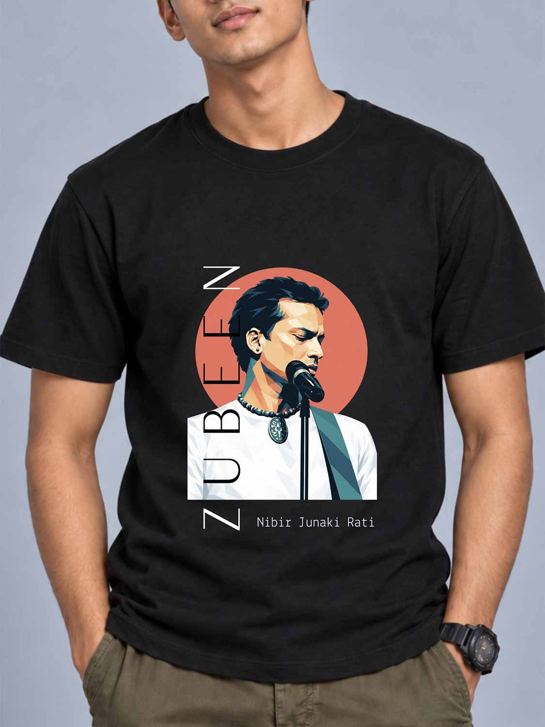 A man wearing a black graphic t-shirt featuring a stylized portrait of Zubeen Nibir Junaki Rati with a red circular background, set against a solid gray studio backdrop.
