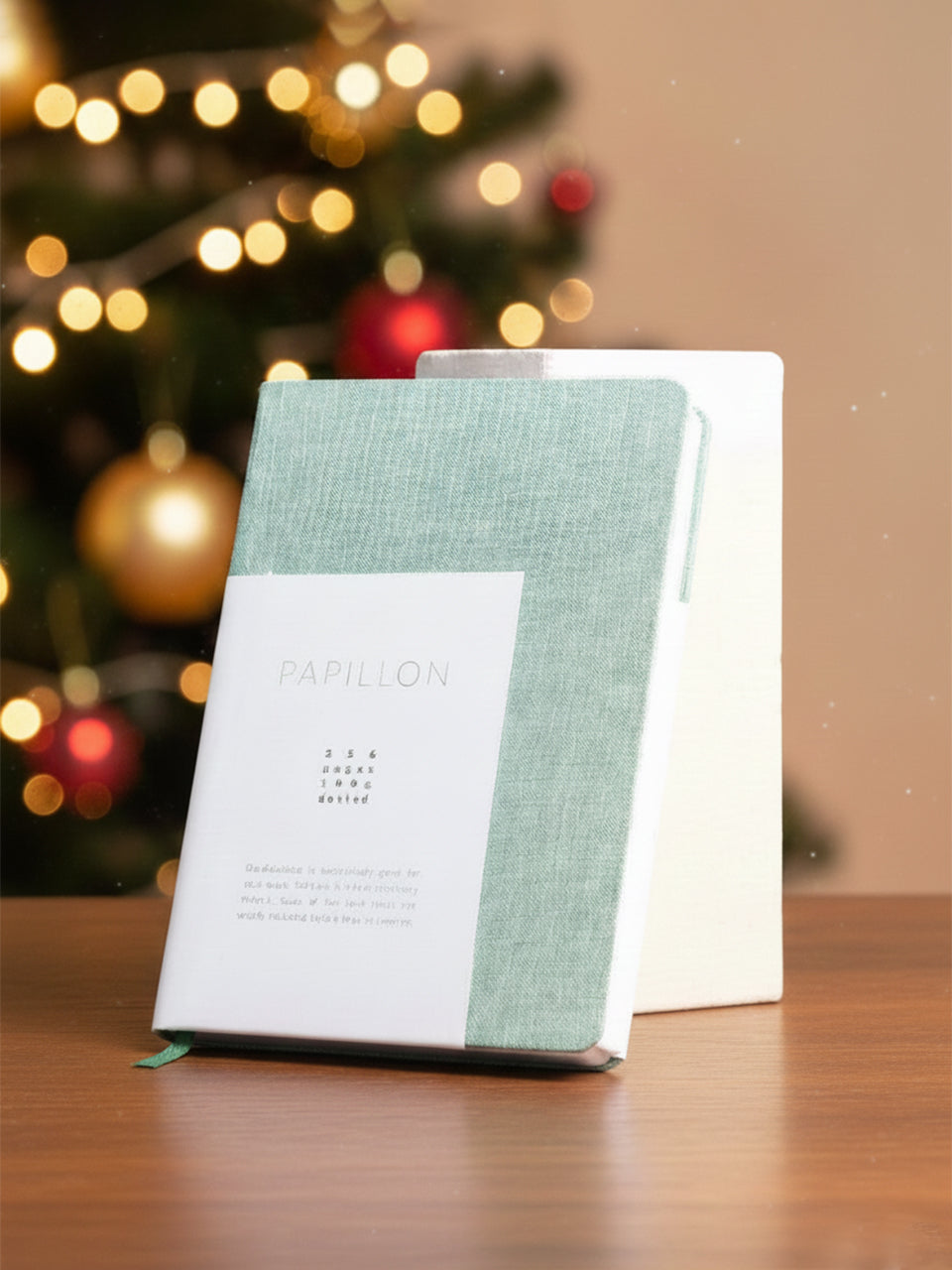Notebook with a green cover and white spine on a wooden surface with a blurred Christmas tree in the background.