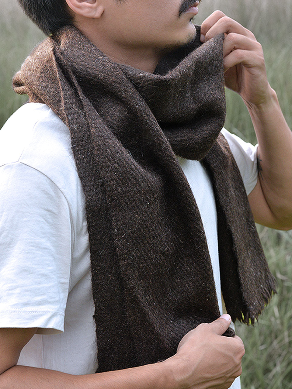 A man wearing a scarf stands in a lush green field, surrounded by nature and open skies