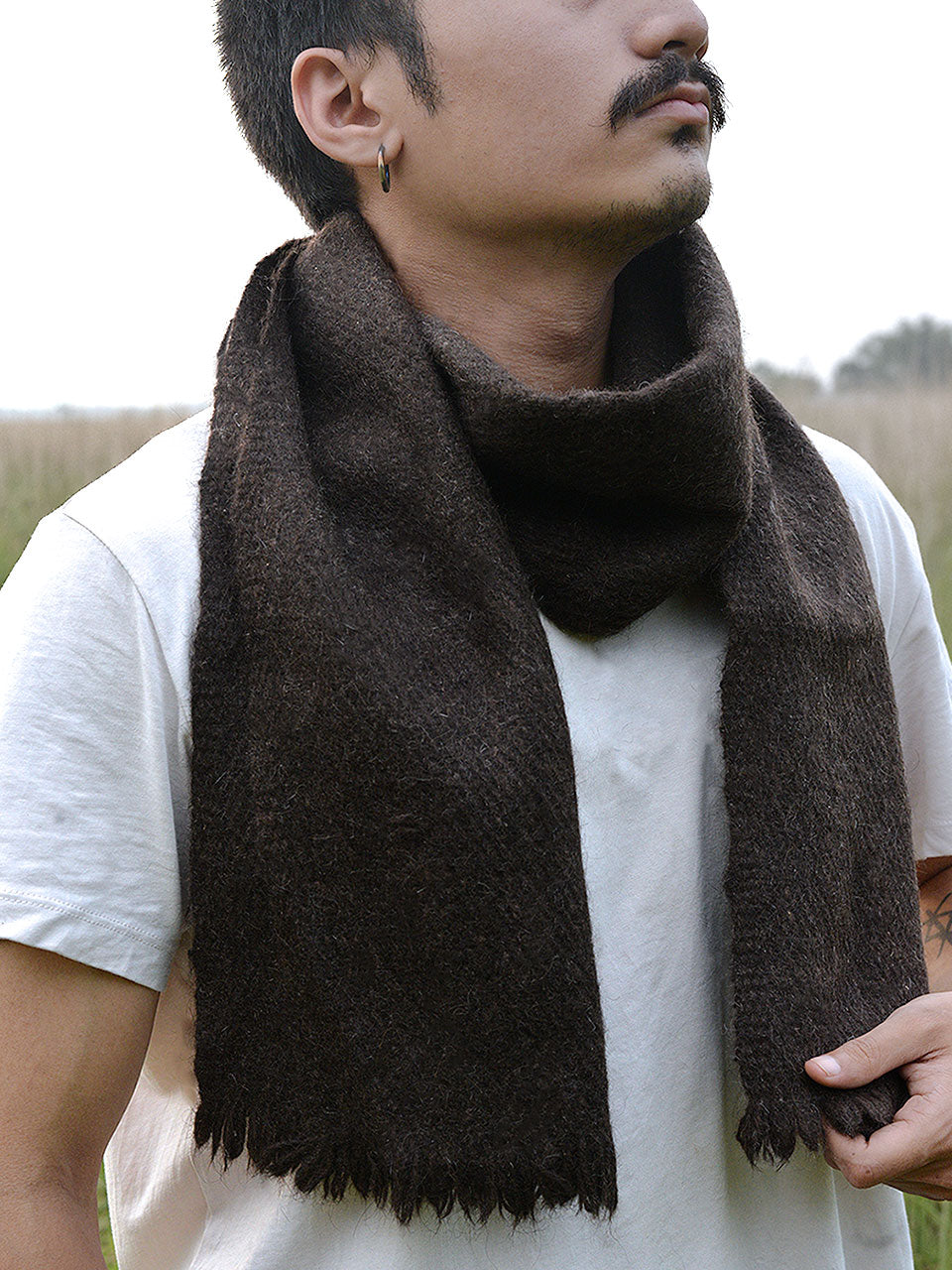 A man wearing a scarf stands in a lush green field, surrounded by nature and open skies