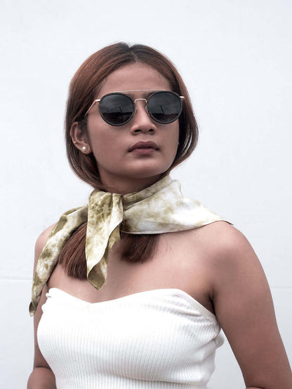 A woman in sunglasses and a white top sits gracefully on a wooden chair with a green scarf, enjoying her surroundings.