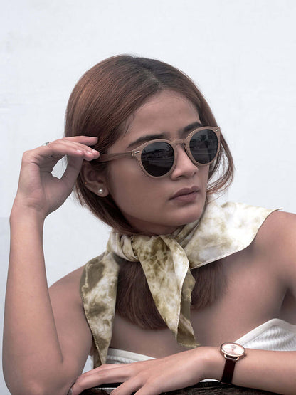 A woman in sunglasses and a white top sits gracefully on a wooden chair with a green scarf, enjoying her surroundings.